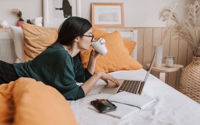 a woman laying on the bed drinking coffee using the laptop